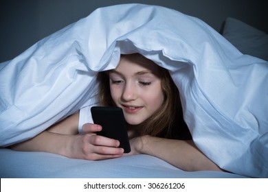 Happy Girl Looking At Mobile Phone While Lying Under Blanket On The Bed