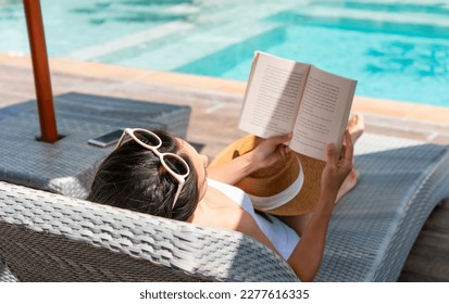 Happy girl lying down on sunbed while reading a book near Swimming Pool on travel holidays vacation, wellness Lifestyle concept. Young Asian woman relaxing at hotel on weekend. Copy space - Powered by Shutterstock