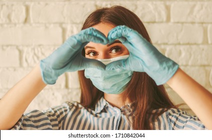Fröhliches Mädchen in medizinischer Gesichtsmaske und Schutzhandschuhe sieht durch Hände in Form von Herzensymbol. Junge Frau in Schutzmaske und Handschuhe, die das Liebeszeichen tragen. Coronavirus COVID-19, Gesundheitswesen