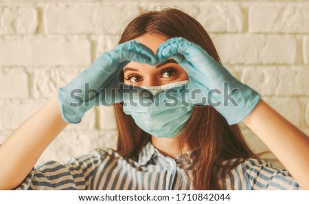 Happy girl in medical face mask and protective gloves looks through hands in shape of heart symbol. Young woman in protective face mask and gloves gesturing love sign. Coronavirus COVID-19, healthcare