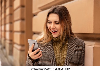 Happy girl with mobile phone in cold weather in street. Young woman using smart phone in winter - Shutterstock ID 1558054850