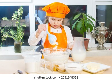 Happy girl in orange chef costume kneads dough with a spoon, child smiles, recipe cooking, girl helps mom cook, child portrait and development.
