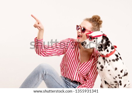 Happy girl and pet in red sunglasses. Young woman hugs dalmatian dog and points on left, isolated on white background. Check this out. Place for text