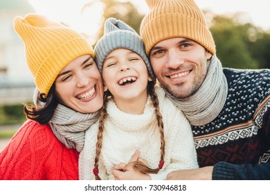 Happy Girl With Pigtails, Wears Warm Knitted Sweater, Stands Between Father And Mother, Laugh Happily, Have Sincere Smiles On Their Faces. Relaxed Family Have Holidays, Spend Nice Time Together