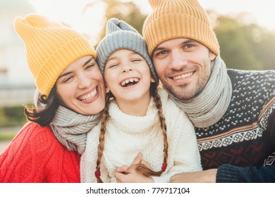 Happy Girl With Pigtails, Wears Warm Knitted Sweater, Stands Between Father And Mother, Laugh Happilly, Have Sincere Smiles On Their Faces. Relaxed Family Have Holidays, Spend Nice Time Together