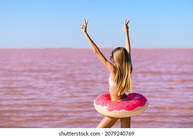 Happy Girl With A Pink Rubber Ring Stands With Raised Hands On The Background Of A Pink Lake.