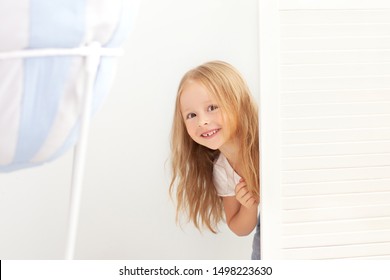 Happy Girl Playing Hide And Seek In Wardrobe. Active Child Looking Hiding Behind A White Door. A Child With A Happy Face, Playing Hide And Seek In The Closet. Positive Baby. Childhood, Fun Concept 