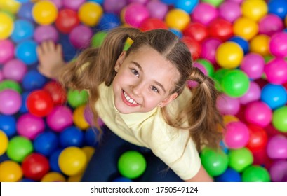 Happy girl with ponytails playing in ball pit at kids playground, top view