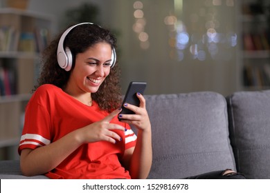 Happy Girl In Red Listening To Music Checking Smart Phone Sitting On A Couch In The Living Room In The Night At Home