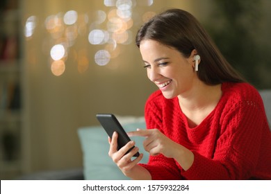 Happy girl in red listening to music with phone and wireless earbuds in the night sitting on a couch in the living room at home