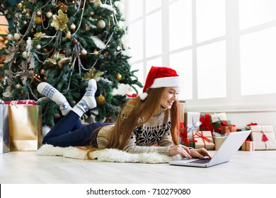 Happy girl in santa hat gereeting friends with christmas in video chat on laptop. Young woman lying under decorated pine tree among gift boxes and packages in home interior with copy space on window - Powered by Shutterstock