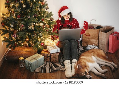happy girl in santa hat shopping online on laptop and sitting with cute dog at golden beautiful christmas tree with lights and presents in festive room. christmas sale and discount concept