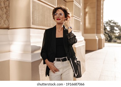 Happy girl with short hair in light trauswrs and dark jacket with handbag smiling outdoors. Modern lady in glasses posing in city..