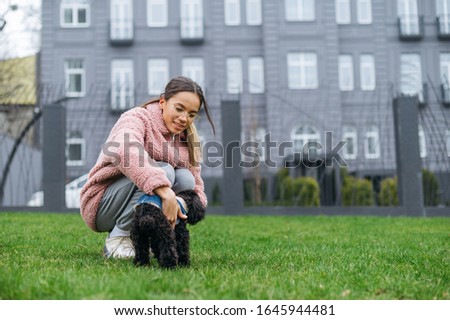 Happy girl sitting on the green lawn and stroking the dog with a smile on her face. Lady in a pink jacket walking a little doggy on the street. Walk with pet concept.
