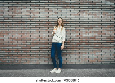 Happy Girl Standing At The Brick Wall Background With Coffee. Urban Fashion Concept. Copy Space