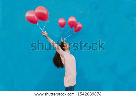 A happy girl stands in front of a blue wall and holds balloons in her hands