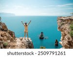 Happy girl stands on a rock high above the sea, wearing a yellow jumpsuit and sporting braided hair, depicting the idea of a summer vacation by the sea.