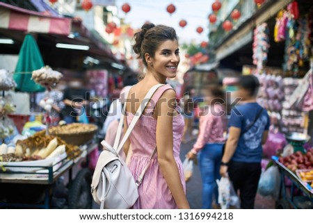 Happy girl in a striped pink dress walks on the asian street market during the celebration of Lunar New Year in Bangkok in Thailand. She has a light backpack and looks into the camera with a smile.