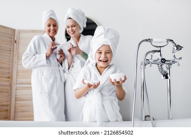 Happy girl in towel and bathrobe holding cosmetic cream near blurred mothers in bathroom