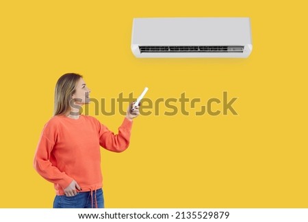 Happy girl turns on the AC. Young lady switches on the AC on the wall. Beautiful woman standing isolated on a yellow colour background setting comfortable temperature her air conditioner at home