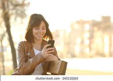 Happy girl using a smart phone in a city park sitting on a bench