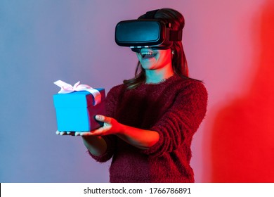 Happy Girl In VR Glasses Holding Wrapped Gift Box And Smiling Joyfully, Giving Christmas Present To Virtual Friend, Celebrating Holidays Birthday In Augmented Reality. Colorful Neon Light Studio Shot