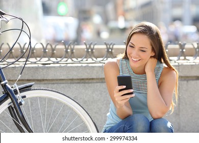 Happy girl watching media in a smart phone sitting in the street beside her bicycle