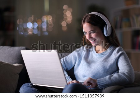 Happy girl wearing headphones watching and listening videos on laptop sitting on a couch in the night at home