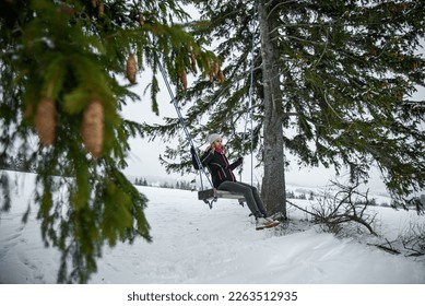Happy girl whit nice smile in dark clothes and a pink hat with a bobble, sitting on a swing between two coniferous trees on a snowy hill in a wild forest. - Powered by Shutterstock