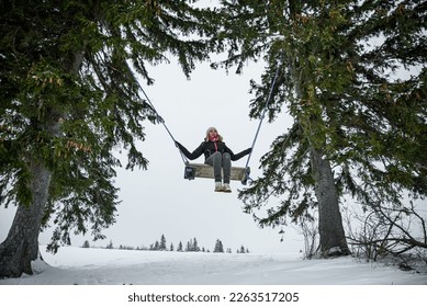 Happy girl whit nice smile in dark clothes and a pink hat with a bobble, sitting on a swing between two coniferous trees on a snowy hill in a wild forest. - Powered by Shutterstock
