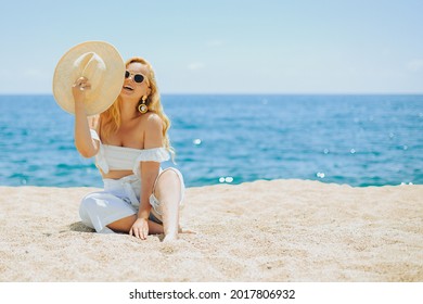 Happy girl in white outfit sits on sand against the backdrop of the sea or ocean beach. Woman smiles and laughs, vacation and joy. Fashion model, beautiful jewelry, earrings, hat, sun glasses.
