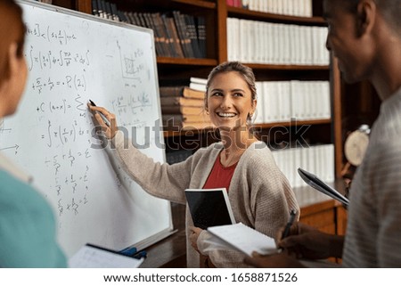 Happy girl writing math formulas on whiteboard. Young woman solving arithmetic problem with university students in classroom. Smiling college student explaining math problem at school.