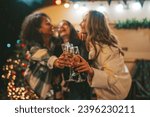 Happy girls celebrating Christmas and New Year winter holidays season outdoor. Young diverse women joyful female friends spending time together have fun drinking sparkling wine near old xmas trailer