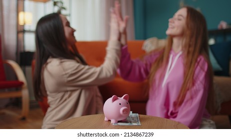 Happy girls friends siblings sitting on floor and take turns dropping dollar banknote into piggy bank, save pocket cash for fun, dreams, future vacation. Keep money, thinking about tomorrow concept - Powered by Shutterstock