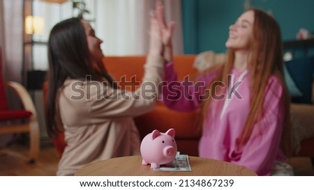 Happy girls friends siblings sitting on floor and take turns dropping dollar banknote into piggy bank, save pocket cash for fun, dreams, future vacation. Keep money, thinking about tomorrow concept