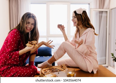 Happy Girls Sitting In Light Flat And Enjoying Fast Food. Glad Sisters Wears Pajamas Joking While Eating Pizza.