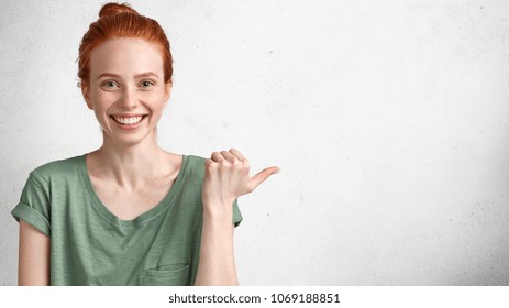 Happy glad beautiful redhead young woman with broad warm shining smile indicates at blank copy space, being satisfied to advertise something, attracts your atention. People, advertisment concept