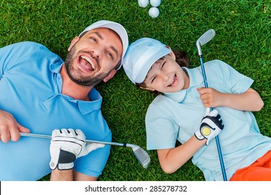 Happy golfers. Top view of cheerful little boy and his father holding golf clubs and smiling while lying on the green grass 