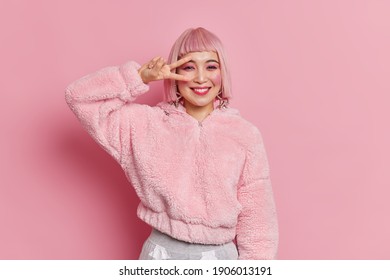 Happy Good Looking Asian Woman With Pink Hair Makes Victory Sign Over Eye Smiles Positively Dressed In Winter Fur Coat Expresses Positive Emotions Poses Indoor. Body Language Concept. Fashion