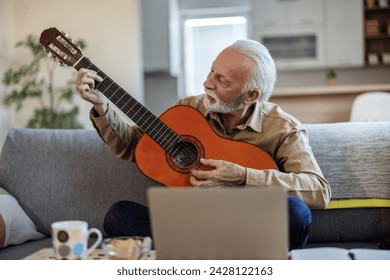 Happy good looking elderly man in a plaid shirt sits on a sofa in the living room and learns to play acoustic guitar online using a Laptop. Senior man learns to play acoustic guitar online. - Powered by Shutterstock