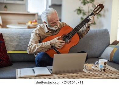 Happy good looking elderly man in a plaid shirt sits on a sofa in the living room and learns to play acoustic guitar online using a Laptop. Senior man learns to play acoustic guitar online. - Powered by Shutterstock