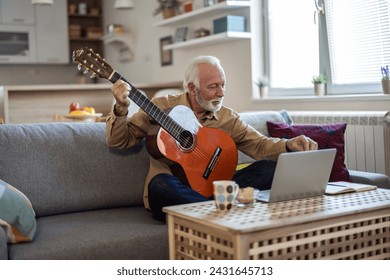 Happy good looking elderly man in a plaid shirt sits on a sofa in the living room and learns to play acoustic guitar online using a Laptop. Senior man learns to play acoustic guitar online. - Powered by Shutterstock