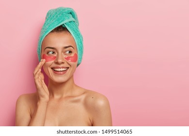 Happy good looking young woman touches gently face, smiles pleasantly, spends free time during weekend on beauty treatments, hydrates delicate skin under eyes, cares about body and complexion