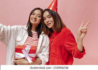 Happy Good-humored Women In Oversized Shirts Take Selfie, Show Peace Sign, Pose With Gift Box And Celebrate Birthday On Pink Background.