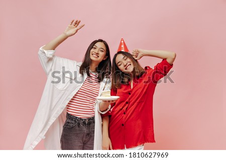 Happy good-humored women in oversized stylish shirts smile and celebrate birthday on pink isolated background. Asian brunette woman holds piece of cake. Young girl poses in party hat.