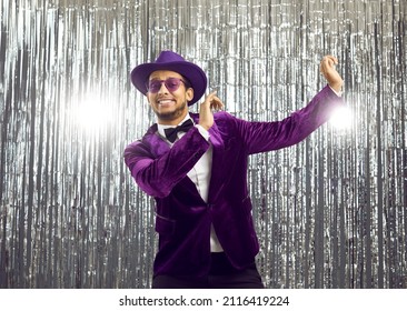 Happy goofy young man wearing a ridiculous tacky unfashionable purple velvet jacket and hat doing funny dance moves at a party. Hilarious dorky guy dancing to music on a silver foil fringe background