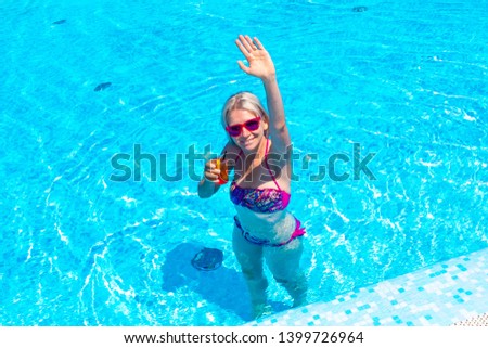  Happy gorgeous female with stylish sunglasses and bikini drinking cocktail and relaxing in the swimming pool at resort spa