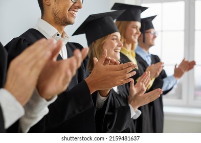 Happy Graduates In Hats And Robes Standing In Row And Applauding. Cheerful Male And Female University Or College Students Having Fun At Graduation Ceremony. Clapping Hands In Soft Selective Focus