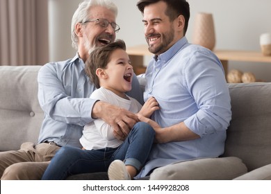 Happy grandfather and father tickling little boy, three generations of men having fun together, sitting on couch, laughing grandparent with son and grandchild relaxing spending weekend at home