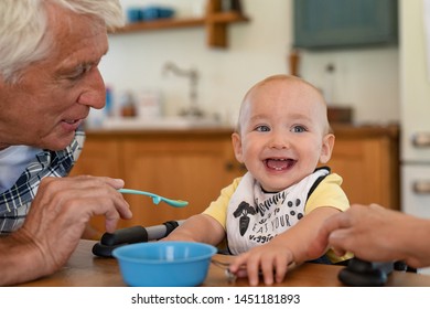 Happy Grandfather Feeding Liquid Food To Grandson Wearing Bib. Cute Adorable Toddler Eating Food With Senior Man. Funny Little Boy Looking Away While Man Feeding With Healhty Fruit Puree, Baby Food.